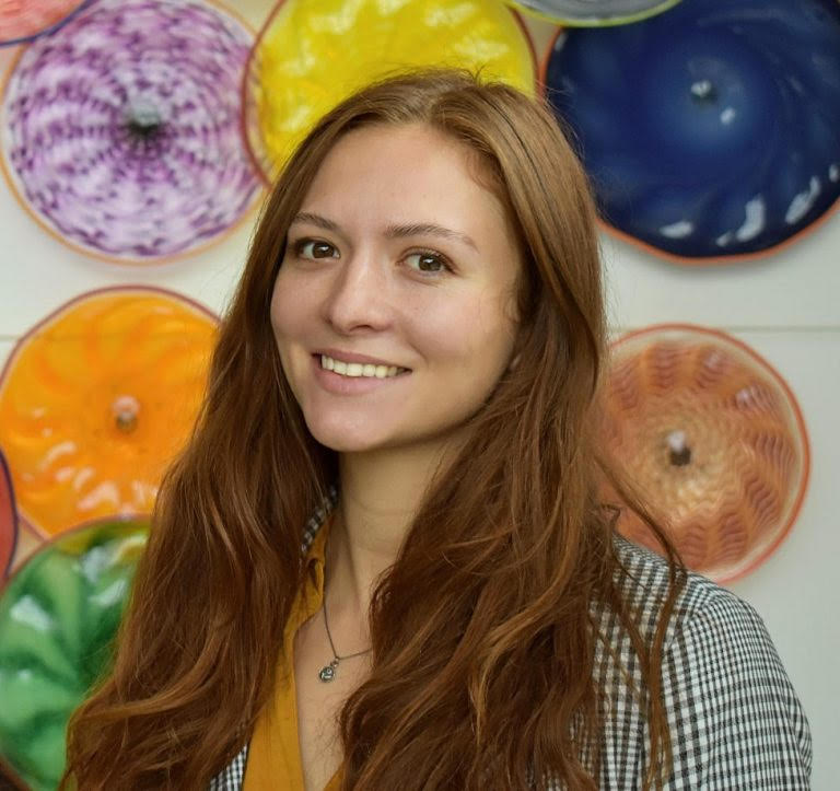 A smiling white woman with red hair and brown eyes, wearing a mustard shirt and a checkered blazer