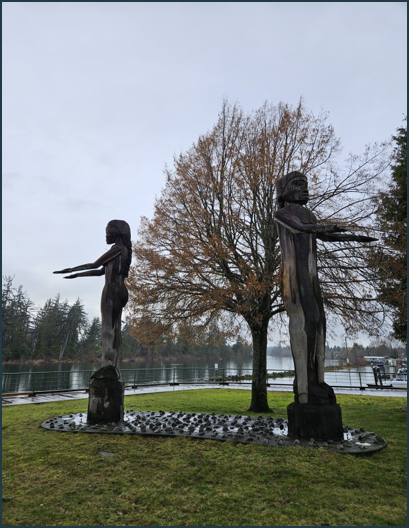 Two tall, wooden figures (male and female) facing away from each other. Their arms are stretched out in front with palms up.